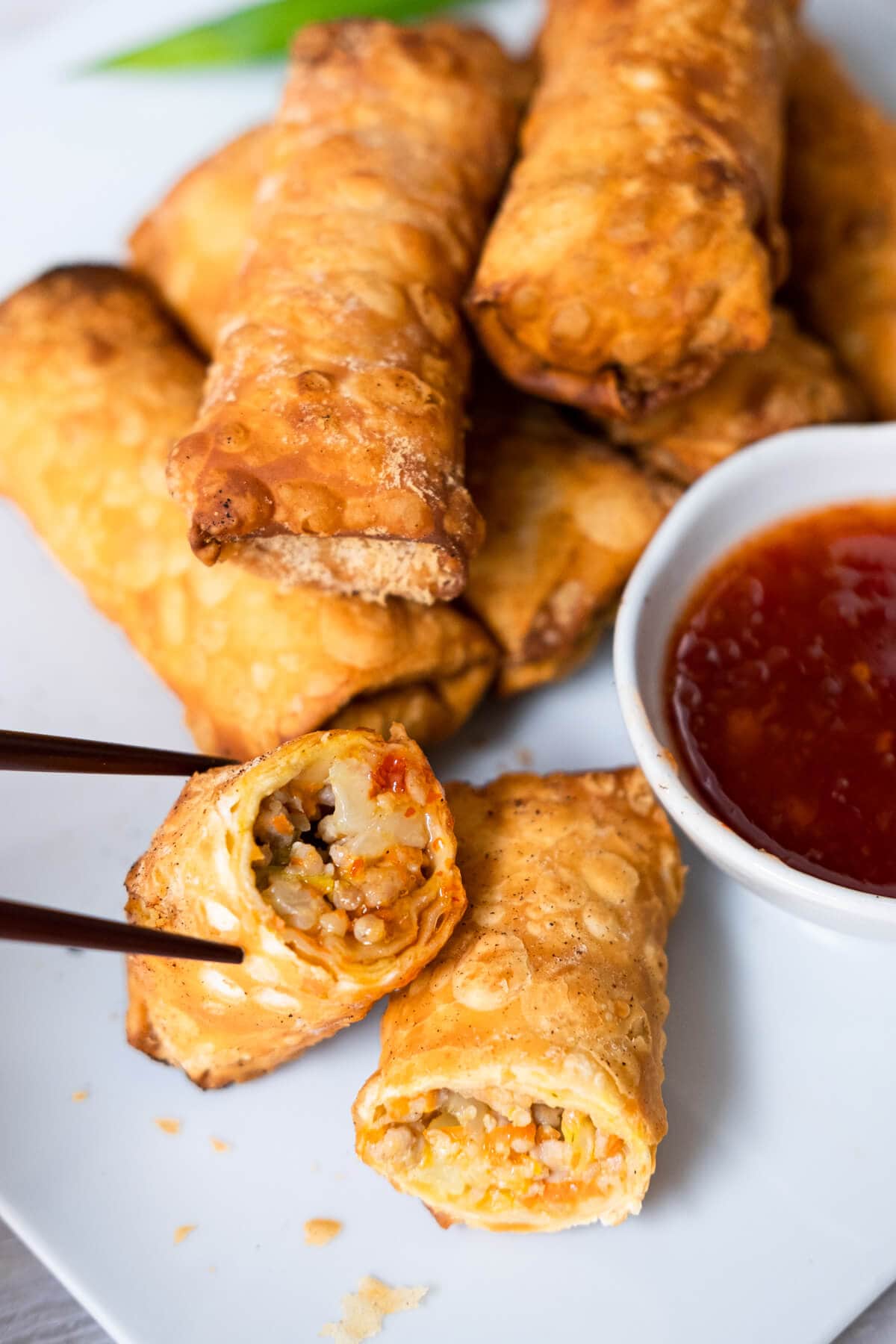 A half cut egg roll picked up with chopstick showing its chicken and vegetables filling with rolls of egg rolls behind it and a small plate of sweet sauce beside. 