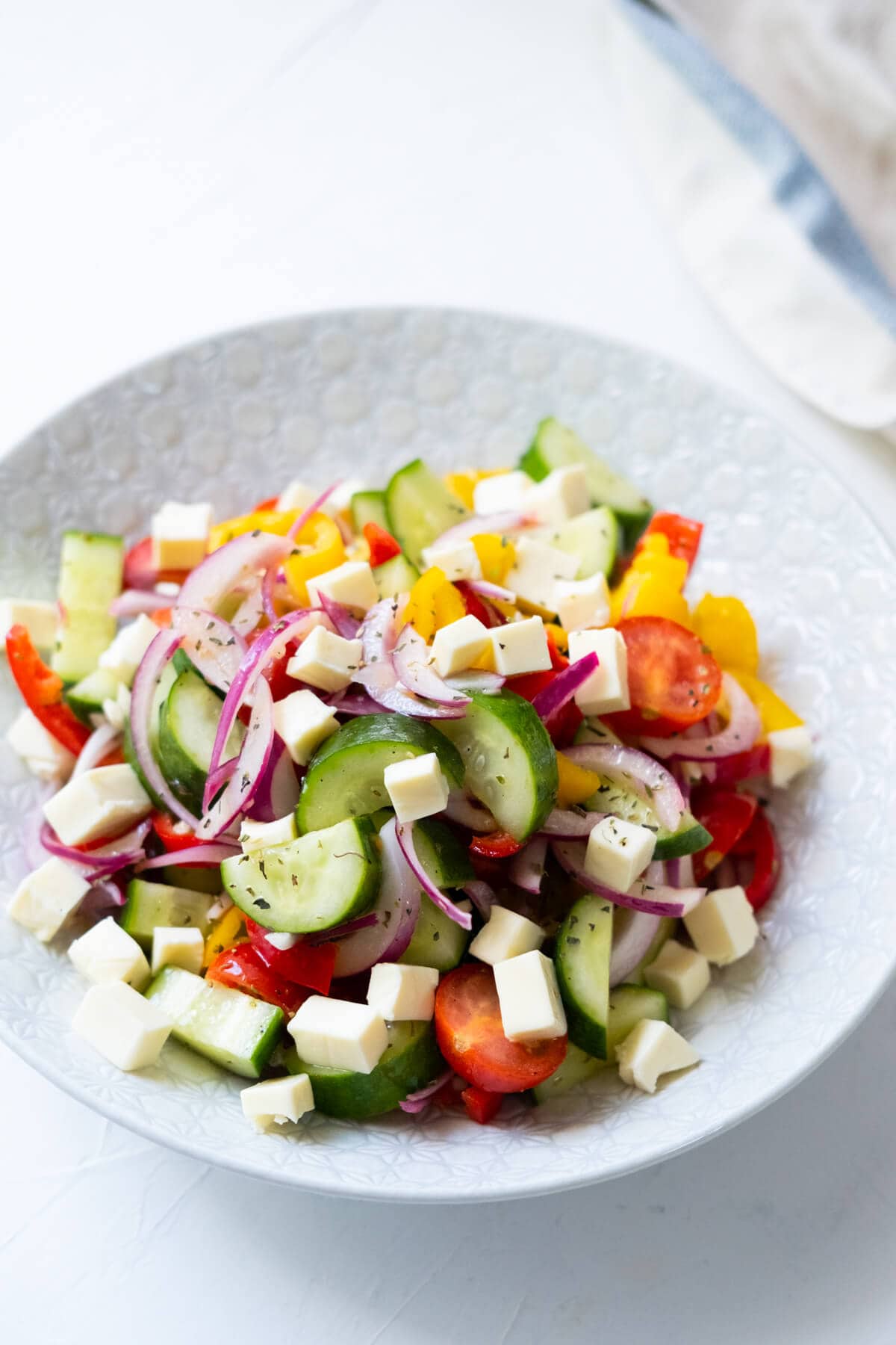 Salad consist of fresh cucumber pieces, red bell pepper pieces, yellow bell pepper pieces, cherry tomatoes, red onions, and feta cheese in a white shallow bowl. 