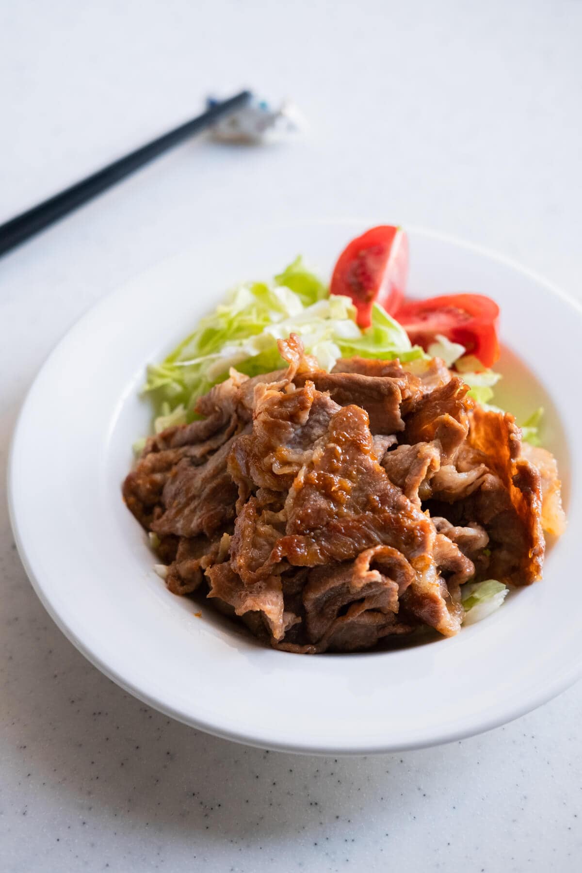 Tender pork slices coated with sweet ginger sauce served with shredded lettuce and sliced tomatoes in a plate with a pair of chopstick on the side.