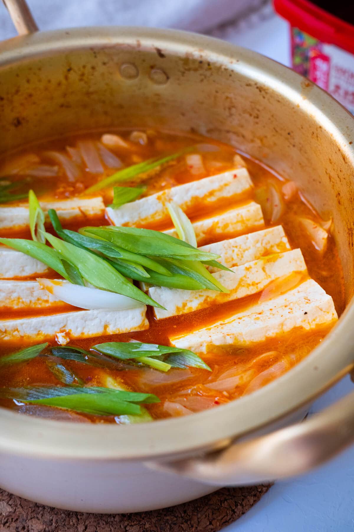 Red kimchi stew with firm tofu slices, pork belly and onion slices served in a metal pot with handles.