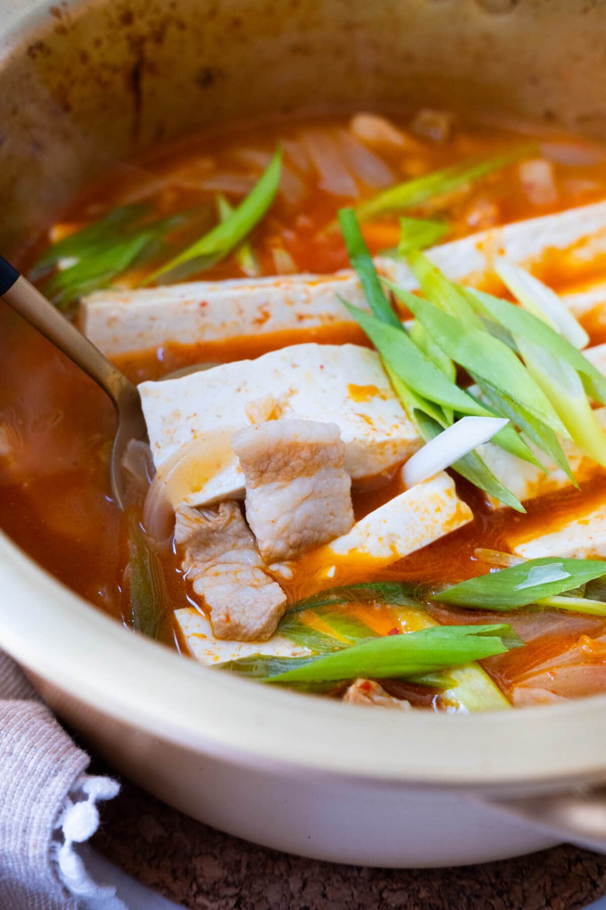 A close up shot of a spoon scooping up sliced tofu and pork belly from a pot of kimchi jjigae. 