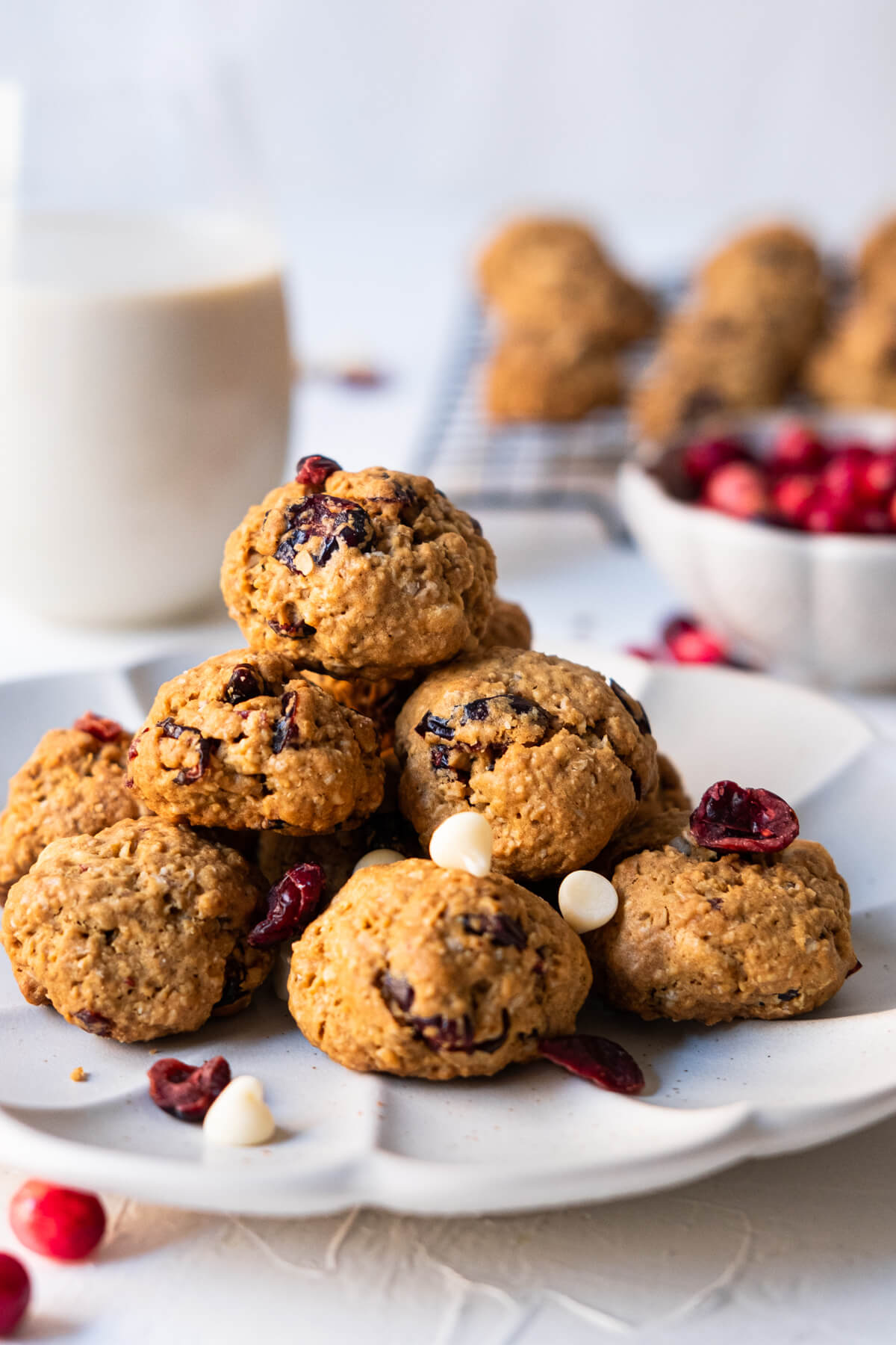Cookies filled with chocolate and cranberry stacked onto each other on a plate with a cup of milk and a bowl of fresh cranberries placed aside. 