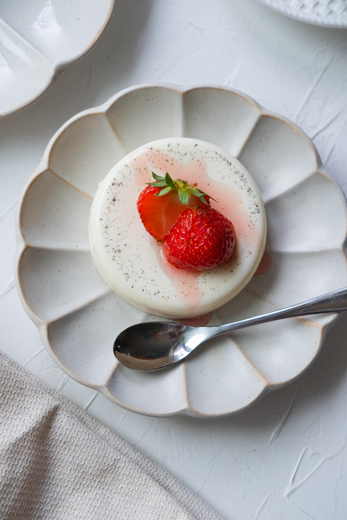 Panna cotta topped with sweet strawberry slices served on a white shallow plate with a spoon aside. 