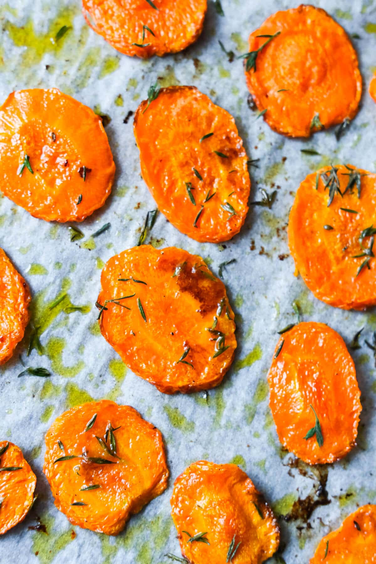 Close up shot of orange baked carrots sprinkled with thyme and olive oil on a baking pan lined with parchment paper.